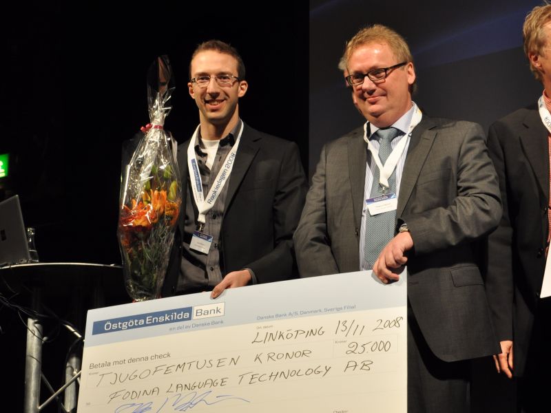 Two men (Lars and Magnus from Fodina) are on a stage. They are holding a bouquet of flowers and a big check for 25,000 Swedish crowns.
